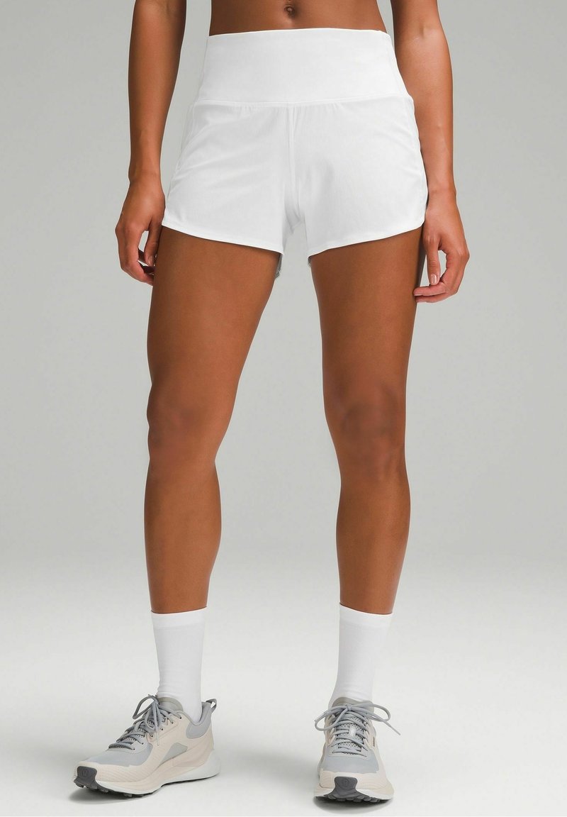 Femme portant un short de sport blanc taille haute, des chaussettes blanches et des chaussures de course grises, debout devant un fond neutre.