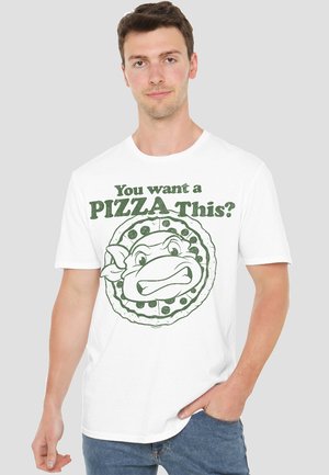 Young man wearing white t-shirt with green graphic of an angry cartoon turtle inside a pizza and text "You want a PIZZA this?"