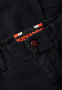 Pantalones azul oscuro con una etiqueta de marca "Superdry" en naranja y blanco visible y un botón marrón grabado con el nombre de la marca.