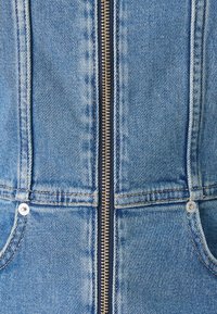 Jeans kombinezon z zadrgo na sprednji strani, svetlo modre barve, z dvema stranskima žepoma in kovinsko opremo. Ima klasično šivanje in teksturirano blago.
