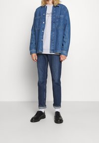 Camisa de mezclilla con dos bolsillos frontales, combinada con un sudadera gris, jeans azules y zapatos negros. Se aprecia una textura de mezclilla de un azul claro.