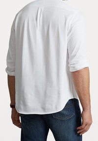 Man draagt een wit poloshirt met lange mouwen opgestroopt en een blauwe spijkerbroek, gezien van achteren tegen een effen achtergrond.