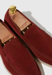 Czerwone zamszowe buty typu slip-on o gładkim wykończeniu, biała podeszwa i drewniane wkładki do butów z złotym pokrętłem. Wnętrze zawiera grawer "Scarosso Made in Italy".