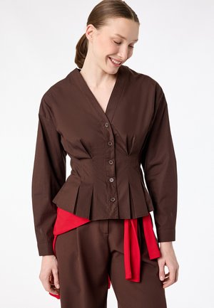 Kobieta ubrana w dopasowaną brązową bluzkę na guziki z plisami oraz pasujące brązowe spodnie, dopełnione czerwonym materiałowym paskiem.