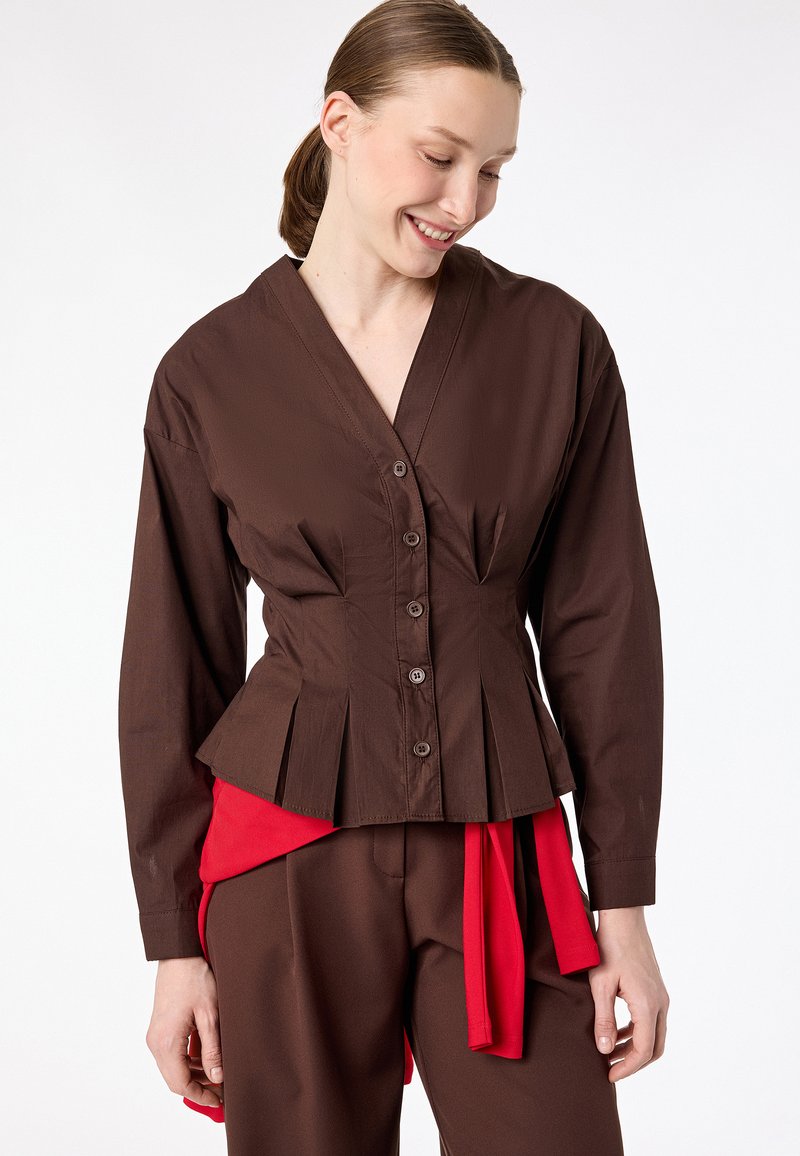Kobieta ubrana w dopasowaną brązową bluzkę na guziki z plisami oraz pasujące brązowe spodnie, dopełnione czerwonym materiałowym paskiem.