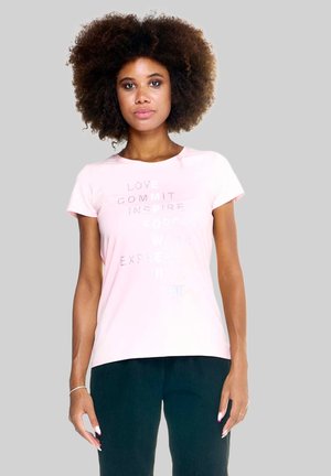 Femme avec coiffure afro portant un t-shirt rose clair avec un texte pâle et un pantalon noir, debout devant un fond gris clair.