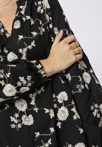 Blusa nera con ricami floreali bianchi, caratterizzata da una vestibilità comoda, maniche lunghe e polsini elasticizzati. Dettagli notevoli includono motivi intricati.