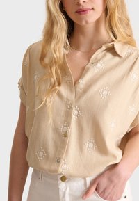 Chemise beige clair à manches courtes et col. Présente des broderies blanches complexes dans des motifs floraux et des fermetures à boutons.