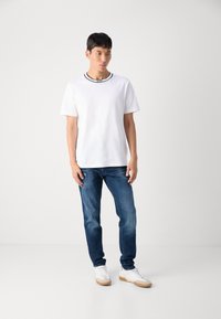 Tricou alb din bumbac cu margini negre și logo, asortat cu blugi din denim blue și teniși albi cu talpă din cauciuc. Design simplu și casual.
