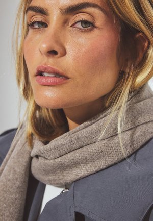 Lähivõte heledate blonde juustega naisest, kes kannab pehmet pruuni kudumist salli ja sinist jakki, millel on sujuvad tekstuurid ja neutraalsed toonid.