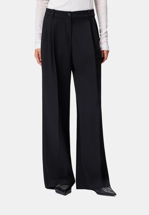 Pantaloni neri a gamba larga a vita alta con pieghe frontali, chiusura con bottone e zip, indossati con una maglia leggera a maniche lunghe e scarpe nere con punte borchiate.