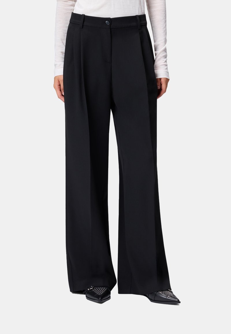 Pantaloni neri a gamba larga a vita alta con pieghe frontali, chiusura con bottone e zip, indossati con una maglia leggera a maniche lunghe e scarpe nere con punte borchiate.