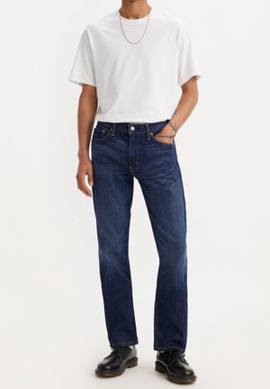 Modèle masculin portant un jean bleu foncé, un t-shirt blanc à manches courtes, des chaussures noires, une chaîne en or, avec une main dans la poche du jean et des tatouages visibles.
