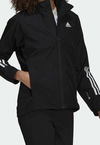 Giacca a vento nera con zip frontale, caratterizzata da strisce bianche Adidas sulle maniche e logo sul petto, realizzata in materiale liscio e leggero.