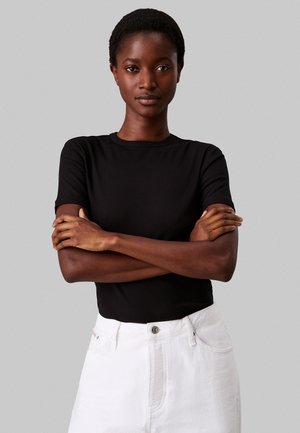 Sort ribbet kortærmet skjorte med rund hals; model iført den med hvide høj taljede jeans, arme korslagt foran.