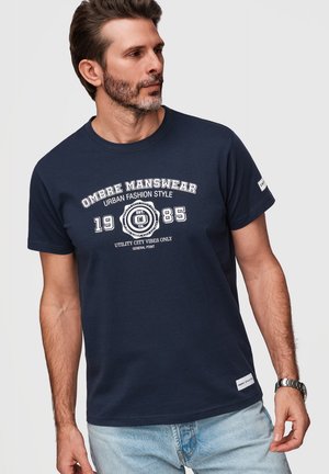 Homme portant un t-shirt graphique bleu marine avec le texte « OMBRE MANSWEAR 1985 Urban Fashion Style », un jean bleu clair et une montre-bracelet argentée.