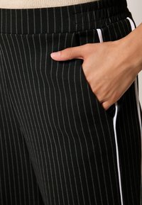 Schwarz gestreifte Hosen mit glatter Textur, die vertikale weiße Nadelstreifen, Seitentaschen und einen elastischen Bund aufweisen.