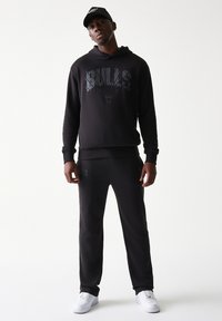 Μαύρο φούτερ και αντίστοιχο παντελόνι φόρμας με τη λέξη "BULLS" σε γκρι χρώμα στο στήθος. Το σύνολο ολοκληρώνεται με λευκά αθλητικά παπούτσια.