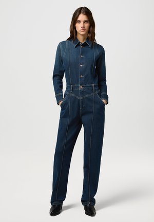 Combinaison en denim avec col, manches longues, boutons sur le devant, poches latérales et coutures contrastées. Couleur bleu foncé avec une texture lisse.