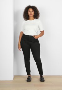 Top bianco a maniche corte abbinato a jeans skinny neri a vita alta e scarpe slip-on nere con dettagli metallici, su uno sfondo semplice.