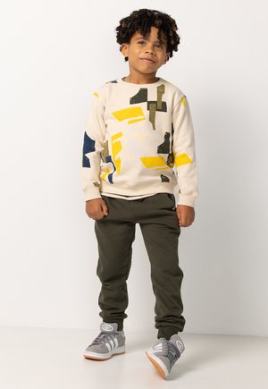 Crème sweatshirt met kleurrijke abstracte patronen in geel, blauw en groen, gecombineerd met olijfgroene broek en grijze sneakers.