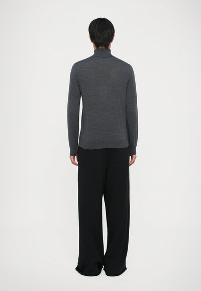 Mann, der mit dem Rücken zur Kamera steht und einen taillierten grauen Rollkragenpullover sowie lockere schwarze weite Hosen trägt, vor einem schlichten Hintergrund.