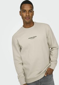 Beige sweatshirt med rund hals, ribbede ærmer og kant, med et sort trykt logo på fronten. Blødt stof, afslappet pasform.