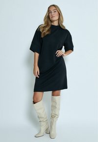 Zwarte oversized jurk met een hoge hals en korte mouwen, gecombineerd met crèmekleurige slouchy kniehoge laarzen met een blokhak.