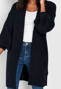 Cardigan tricoté bleu marine foncé à ouverture avant, manches larges, texture côtelée, coupe décontractée, porté sur un haut blanc et un jean bleu.