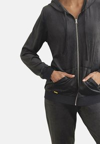 Zwarte zip-up hoodie van zacht materiaal; heeft een capuchon, twee voorkers, en ribbels aan de mouwen en onderkant.