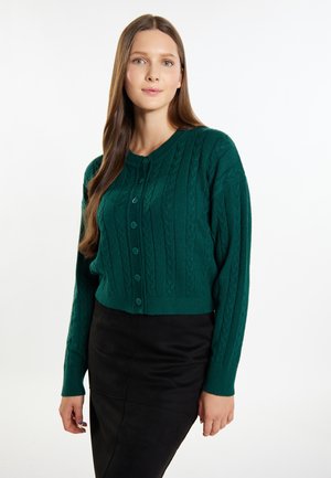 Grüne Strickjacke mit einem Zopfmuster, Rundhalsausschnitt und Knopfleiste. Wird über einem schwarzen Rock getragen, hat lange Ärmel und einen verkürzten Schnitt.