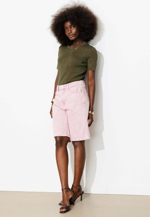 Grande femme avec coiffure afro portant un haut vert olive à manches courtes, un short rose pâle à hauteur des genoux, des sandales noires à talons et des bijoux en or.