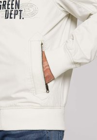 White bomber jacket with a quilted texture, featuring a zippered pocket and ribbed cuffs, highlighted by dark graphic text on the front.