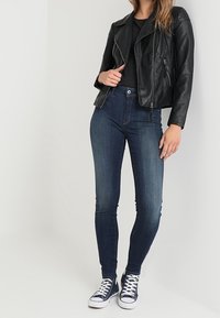Chaqueta de cuero negra de motorista sobre una camiseta negra, combinada con jeans ajustados azul oscuro y zapatillas navy con detalles blancos.