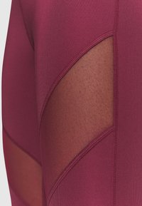 Leggings burdeos con paneles de malla. La tela es suave, con un diseño ajustado y acentos transparentes en los laterales.