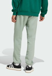 Pantaloni de trening verde deschis cu manșete elastice și cusături laterale, asociați cu adidași negri Adidas, având dungi și logo alb.