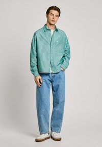 Camicia a righe verdi con un taglio rilassato, abbinata a jeans larghi di colore azzurro chiaro e sneakers bianche con suola marrone.