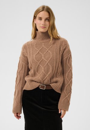 Femme aux cheveux blonds mi-longs portant un pull à col roulé en maille torsadée marron et une jupe noire avec une ceinture marron sur un fond uni.