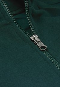 Chaqueta de color verde oscuro con cremallera, que presenta una cremallera metálica con un tirador texturizado sobre un fondo de tela suave.