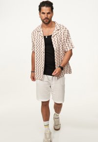 Kurzärmliges Hemd mit Zickzack-Muster in Creme und Maroon über einem schwarzen Tanktop, kombiniert mit weißen Shorts und beigen Sneakers.