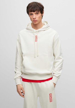 Felpa color crema con tasca frontale, cordini, dettagli in rosso del logo. Indossata con pantaloni jogger abbinati color crema con accenti rossi. Taglio semplice e rilassato.