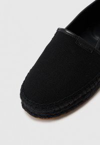 Zapatilla negra de lona sin cordones con un upper texturizado, suela de goma y detalles cosidos a lo largo del borde. Presenta una etiqueta de logo en el costado.