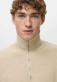 Beige gebreide trui met hoge kraag en halve ritssluiting. Getextureerde stof en aansluitend model, gedragen door een model met krullend haar.