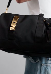 Person wearing light blue jeans carries black duffle bag with gold Moschino logo and black handles over shoulder.