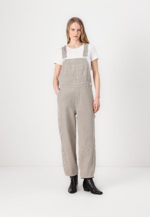Salopette rayée en gris clair et blanc, avec des bretelles réglables, des poches latérales et une coupe ample. Portée avec un t-shirt blanc et des bottes noires.