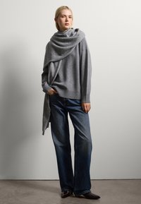 Grauer Strickpullover mit einem drapierten Schalkragen, kombiniert mit dunkelblauen weit geschnittenen Jeans und spitzen braunen Schuhen. Glatte Textur und minimalistisches Design.