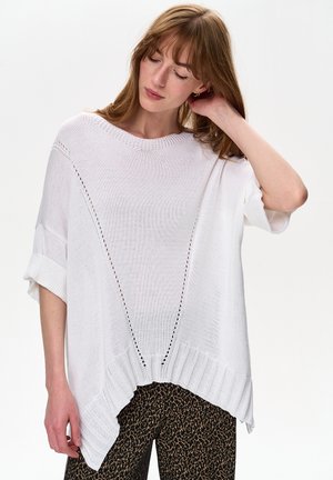 Biały dziergany sweter z okrągłym dekoltem, ozdobiony ażurowymi detalami i ściągaczem na dole. Do tego spodnie w panterkę.