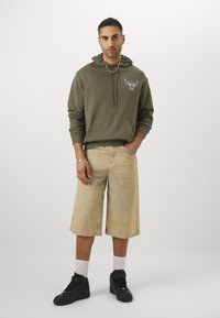 Oliven grøn hoodie med en okse-logo, beige denim shorts, sorte høj-top sneakers og hvide crew-sokker. Casual, afslappet pasform design.