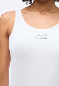 Κοντινό πλάνο ενός ατόμου που φοράει λευκό αμάνικο τοπ με το λογότυπο "EA7 Emporio Armani" στο στήθος.