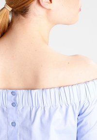 Jasnoniebieska bluzka typu off-the-shoulder z bufkami, z elastycznym marszczeniem przy dekolcie i zapięciem na guzik z tyłu.